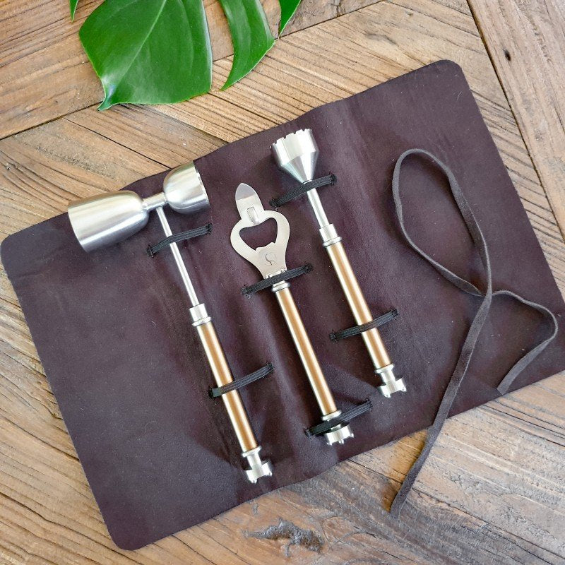 Set of bar tools including a mallet, bottle opener, and corkscrew on a brown leather case with a leaf in the background.