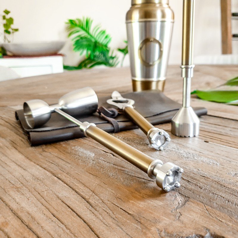 Bar tools including a muddler, jigger, and shaker on a wooden surface with a blurred background.