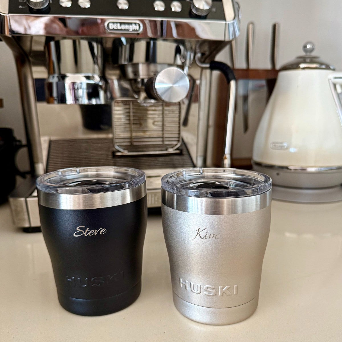 Two insulated coffee cups labeled 'Steve' and 'Kim' in front of a Delonghi espresso machine Huski short tumbler engraved 