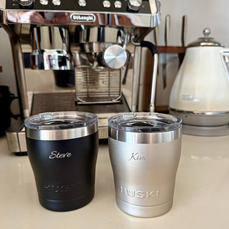 Two insulated coffee cups labeled 'Steve' and 'Kim' in front of a Delonghi espresso machine Huski short tumbler engraved 